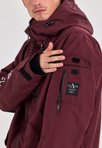 Maroon jacket with a hood, featuring a zippered pocket, Velcro accents, and drawstrings. Textured fabric with branding detail on the arm.