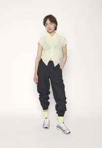 Camisa de botón amarilla clara de manga corta con un patrón texturizado, combinada con pantalones negros de corte holgado y zapatillas blancas con detalles en neón.