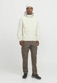 Chaqueta cremosa con cremallera y capucha, puños elásticos y detalles en negro, combinada con pantalones cargo marrones que tienen bolsillos laterales y zapatillas negras.