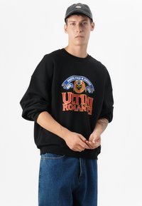 Zwarte sweatshirt met een kleurrafische opdruk met de tekst "ULTIMI ROMANTICI," gecombineerd met blauwe denim jeans en een grijze pet. Neutrale achtergrond.
