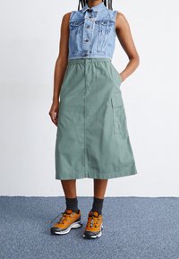 Jeansowa kamizelka bez rękawów z kieszeniami na piersi, zestawiona z zieloną, sięgającą połowy długości spódnicą z elastycznym pasem i boczną kieszenią. Pomarańczowe buty sportowe.