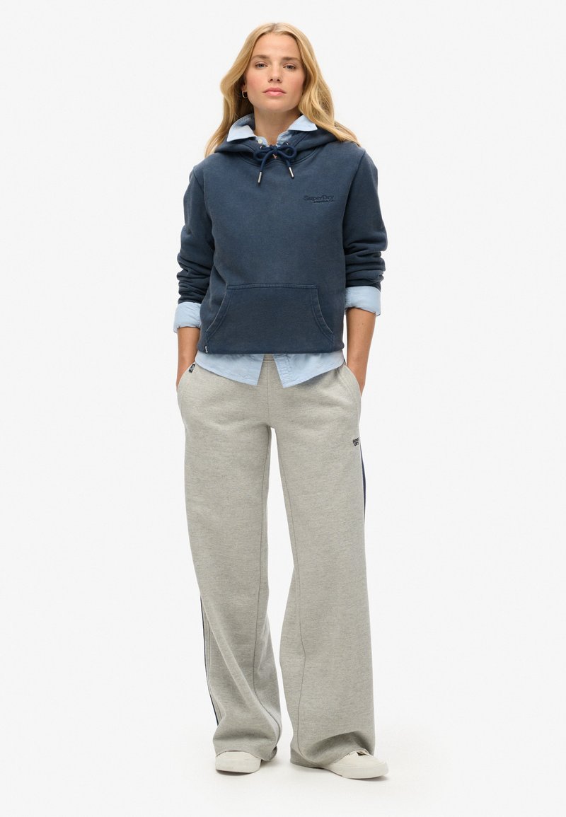 Marineblå hættetrøje med snor, frontlomme, lagdelt over en lyseblå skjorte. Grå sweatpants med sideliner og logo. Hvide sneakers.