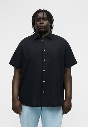 Sötét bőrű, rövid rasztahajú személy laza fekete, rövid ujjú, gombos ingben és világoskék farmerral, semleges pózban áll.