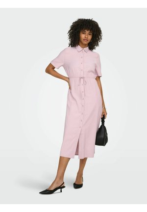 Nainen, jolla on kiharat hiukset, pukeutuneena vaaleanpunaiseen polvipituiseen mekkoon, jossa on napit ja kiristysnauha vyötäröllä, mustat slingback-korkokengät ja musta käsilaukku kädessään.