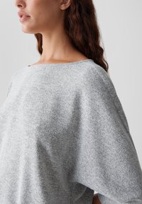 Femme aux longs cheveux bruns portant un pull en laine léger gris clair, vue de côté et du cou jusqu'au milieu du torse, sur un fond uni.
