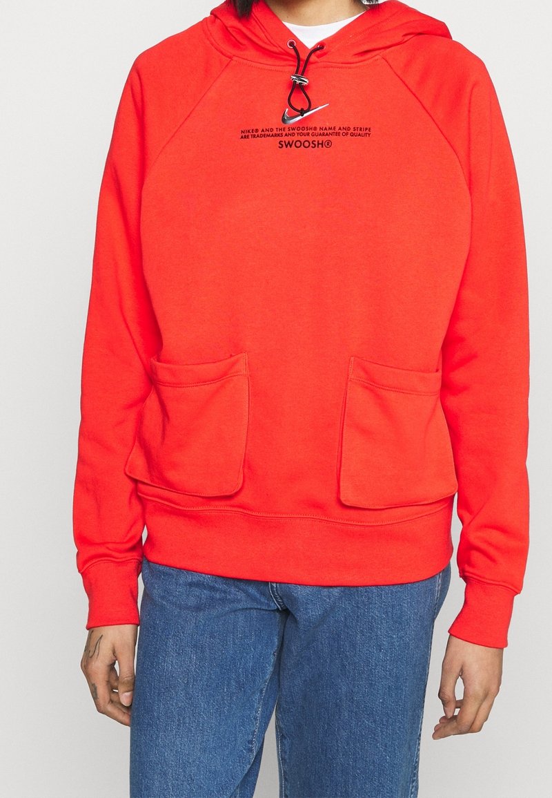 Sweat à capuche rouge vif avec deux poches frontales, cordon noir réglable, et petit texte noir au-dessus de la poitrine indiquant "SWOOSH", porté avec un jean bleu.