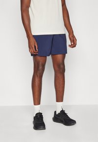 Navy athletic shorts with a smooth texture, elastic waistband, and subtle side slits, paired with white socks and black sports shoes.