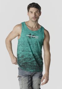 Hombre con camiseta sin mangas turquesa con patrón moteado negro y logo "KOROSHI BRAND", combinado con pantalones cargo de mezclilla clara.