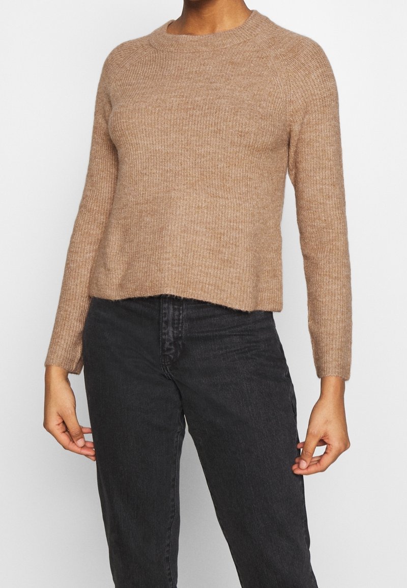 Personne portant un pull en maille côtelée marron clair à manches longues et un jean noir taille haute, debout devant un fond uni.