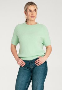 Miętowy sweter z krótkim rękawem o miękkiej fakturze, okrągłym dekolcie i ściągaczu, połączony z ciemnoniebieskimi dżinsami. Minimalistyczny design.