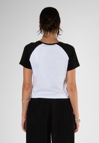 Camiseta de manga corta tipo raglán con mangas negras y cuerpo blanco, hecha de algodón. Presenta un cuello redondo y un ajuste casual, combinada con pantalones negros con plisados.