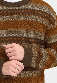 Brązowy i kremowy sweter wzorzysty z okrągłym dekoltem, z teksturowanymi paskami i dzianymi mankietami. Materiał wydaje się miękki i ciepły.