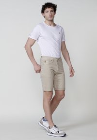 Ung man står i beige shorts, vit t-shirt, mönstrade strumpor och vita sneakers mot en ljus, enhetlig bakgrund.