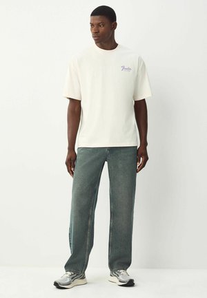 Camiseta de color crema con mangas cortas, que presenta un pequeño logo morado. Combinada con unos vaqueros de corte holgado y textura de color azul claro y zapatillas grises.