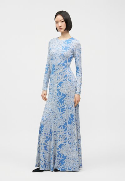 Γυναίκα που φοράει μακρυμάνικο, μακρύ μπλε φόρεμα με λευκά σχέδια φύλλων, στέκεται μπροστά σε απλό ανοιχτό γκρι φόντο.