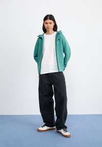 Turquoise zip-up hoodie with black Nike logo, white t-shirt, loose black jeans, and white shoes with brown soles on blue background.