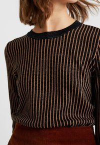 Haut à manches longues rayé noir et or avec une texture côtelée et un col rond, porté avec une jupe en velours côtelé marron.