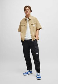 Beige short-sleeve button-up shirt, white T-shirt underneath, black cargo pants, blue and white sneakers, featuring pockets and textured fabrics.