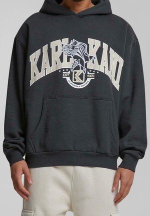 Ciemnoszara bluza z kapturem z dużymi literami "KARL KANI", grafiką orła i napisem "EST. 1989", noszona z jasnymi spodniami.