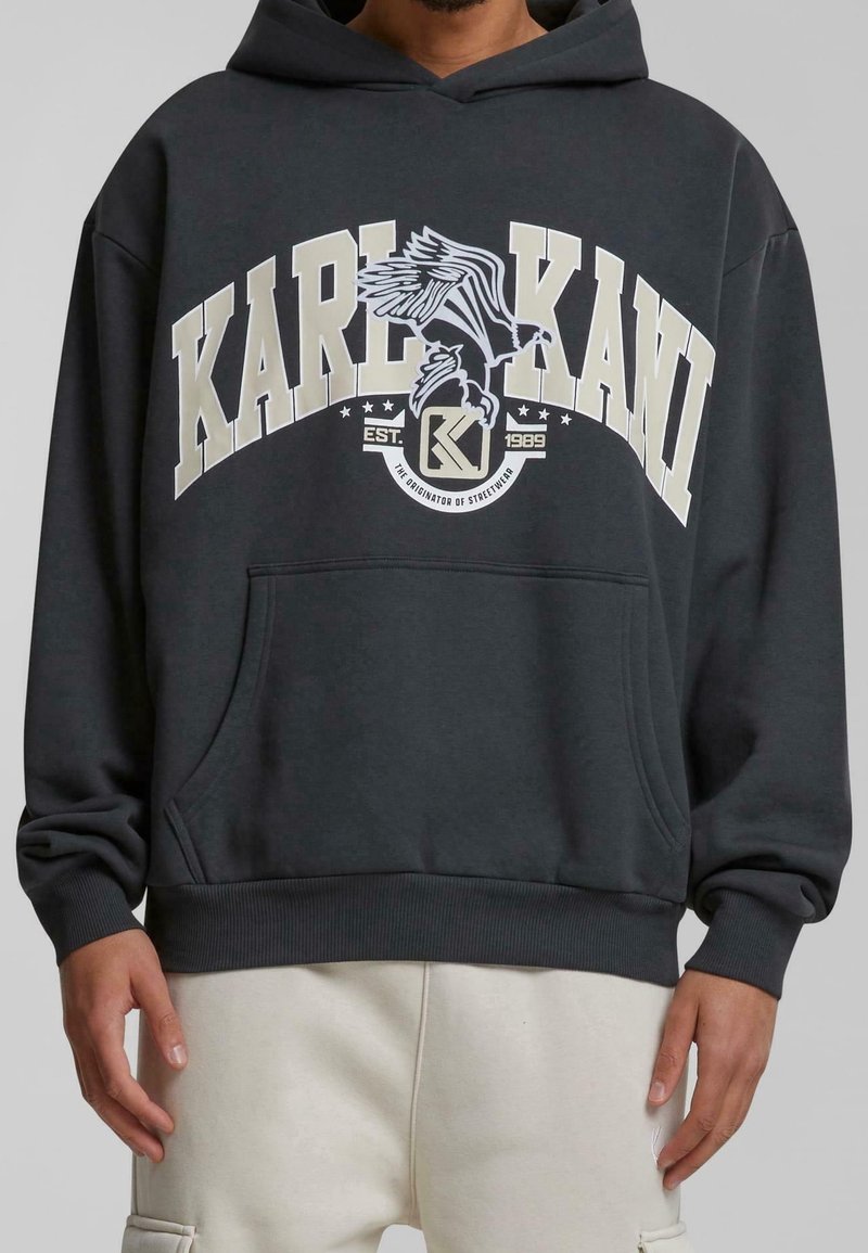 Sweat à capuche gris foncé avec « KARL KANI » en grandes lettres, motif d'aigle et texte « EST. 1989 », porté avec un pantalon clair.