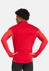 Maglia sportiva rossa a maniche lunghe con colletto alto, design aderente, superficie liscia e pannelli a contrasto su spalle e maniche.