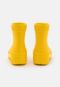 Par de botas de goma amarillas brillantes vistas desde atrás, con puntas texturizadas y suelas gruesas sobre un fondo blanco.