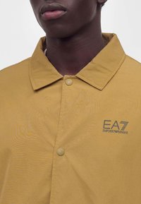 Близък план на човек, носещ горчици жълто яке с яка с лого EA7 Emporio Armani на лявата страна на гърдите, застопорено с копчета.