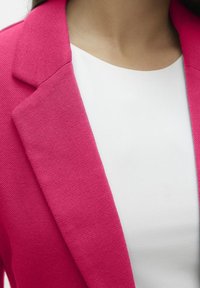 Blazer fucsia con tessuto testurizzato, dotato di rever a punta, abbinato a un top bianco a collo rotondo e liscio sotto.