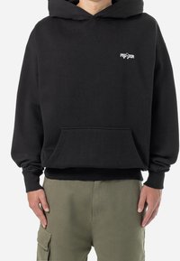 Person, der einen schwarzen Kapuzenpullover mit Fronttasche und weißem "PEQUODOR"-Schriftzug auf der Brust trägt, kombiniert mit olivgrünen Cargo-Hosen.