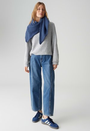 Grijze textuurtrui, blauwe oversized spijkerbroek en blauwe sneakers met witte strepen. Blauwe driehoekige sjaal zorgt voor een gelaagd detail.