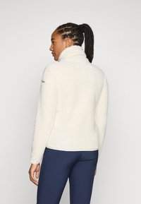 Veste en polaire crème avec un col haut, une surface texturée et une coupe slim. Associée à des leggings de sport bleu foncé.