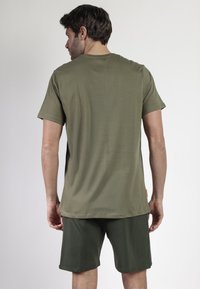 Hombre de pie de espaldas, vistiendo una camiseta de manga corta verde oliva y pantalones cortos verde oscuro, contra un fondo plano.