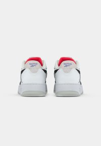Bijele kožne tenisice s crnim akcentima, sivim suedenim detaljima i crvenim unutrašnjim dijelom. Imaju debeo bijeli potplat i ljubičasti Reebok logo.