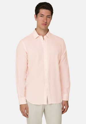 Jonge man die een lichtroze overhemd met lange mouwen en beige broek draagt, staand tegen een effen witte achtergrond.