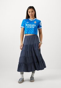 Maglietta sportiva blu corta con strisce bianche e logo, abbinata a una lunga gonna a strati a quadri in blu scuro e bianco. Calzature e calze grigie.