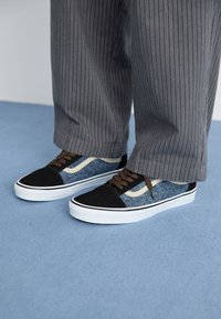 Black and blue sneaker with denim and suede material, white rubber sole, brown laces, and cream accents. Worn with striped gray pants.