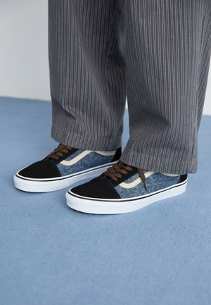 Black and blue sneaker with denim and suede material, white rubber sole, brown laces, and cream accents. Worn with striped gray pants.
