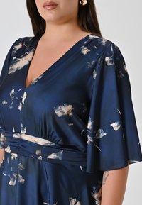 Marineblaues Kleid aus leichtem Stoff mit einem floralen Muster in Creme und Beige, das einen tiefen V-Ausschnitt und geflütterte Ärmel hat.