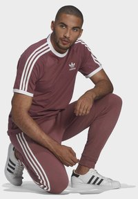 Adidas tumši sarkans sporta uzvalks ar baltām detaļām un trim svītrām uz piedurknēm un biksēm. Stilizēts ar baltām čībām ar melnām svītrām.