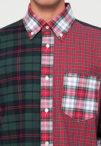 Karierter Hemd mit einer Mischung aus grünem und rotem Plaid-Muster, weißen Knöpfen und einer Brusttasche. Der Kragen ist ebenfalls im Karomuster.