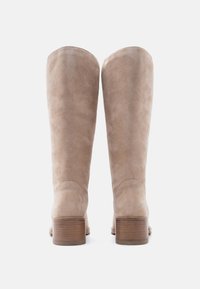 Paire de bottes hautes en daim beige avec talons en bois empilés vues de dos sur fond blanc.