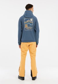 Blaue Kapuzenjacke mit einem grafischen Design, das Berge und Bäume zeigt, kombiniert mit gelben Hosen, die eine lockere Passform und kontrastierende Farben zeigen.