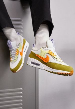 Persona con zapatillas Nike blancas, naranjas y verdes, calcetas blancas y pantalones negros, sentada en un casillero metálico gris.