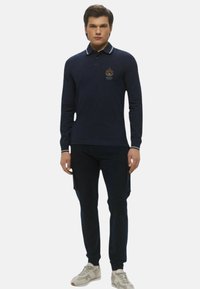 Giovane in piedi, indossa una polo a maniche lunghe blu navy con emblema, pantaloni neri slim e sneakers bianche, sfondo neutro.