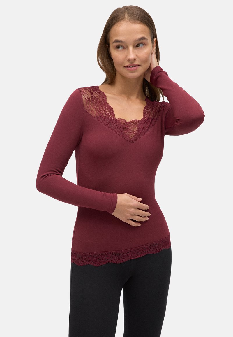 Top burdeos de manga larga con cuello y dobladillo de encaje. Diseño suave y ajustado con una textura lisa, combinado con leggings negros.