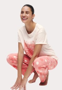 Helles rosa T-Shirt mit Sonnenmotiv und kurzen Ärmeln, kombiniert mit rosa Hosen mit weißen Sonnenstrahlen-Mustern. Das Modell hockt barfuß.
