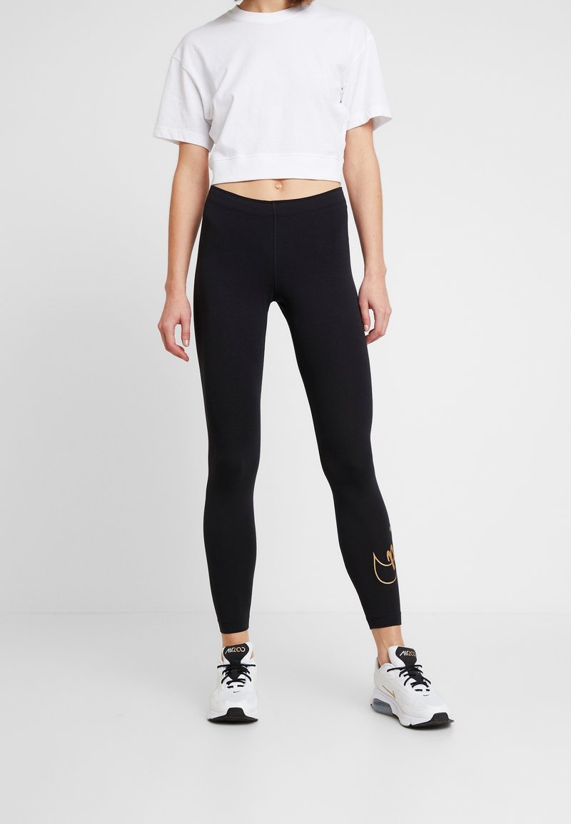 Svarta leggings med en slät textur, prydda med en guldfärgad logotyp på vänstra benet. Kombinerat med en avkortad vit t-shirt och sportskor.