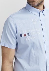 Camisa de manga corta azul claro con cuello abotonado. Presenta texto y logotipos bordados en blanco y rojo. Tejido de algodón suave.