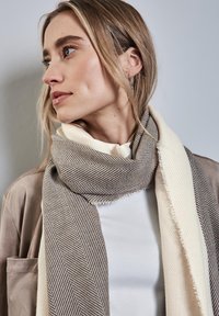 Gewebter Schal mit einem Fischgrätmuster in Braun und Creme, mit weicher Textur und Fransenrändern, um den Hals drapiert.