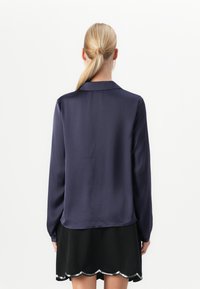 Woman with blonde hair in a low ponytail wears a long-sleeve navy blouse and a black skirt with a silver scalloped hem, facing away.